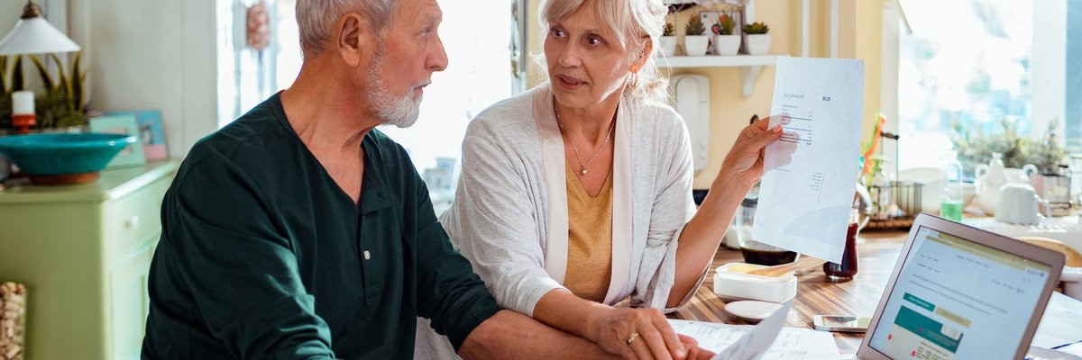 An older couple pays bills at the kitchen table using a laptop.