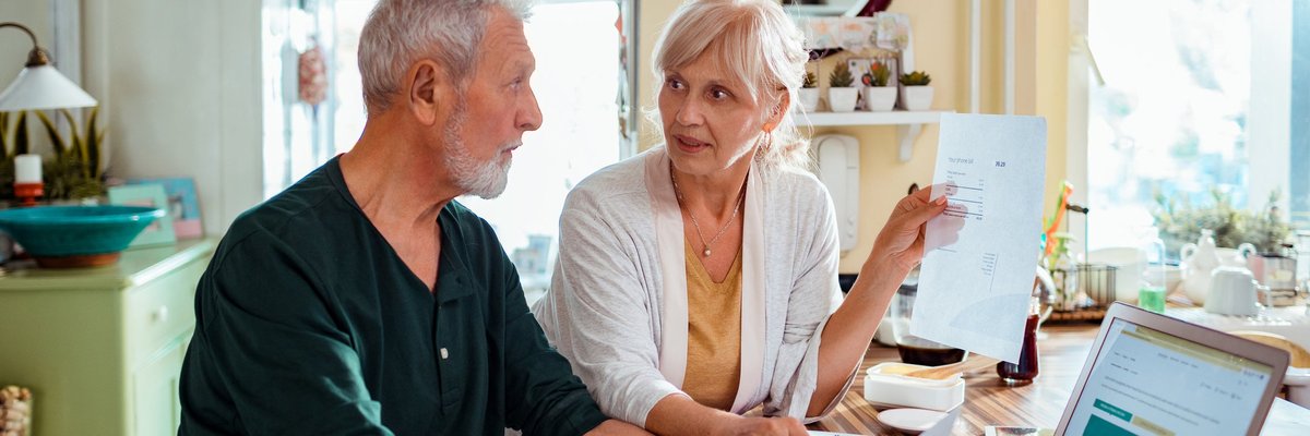 An older couple pays bills at the kitchen table using a laptop.
