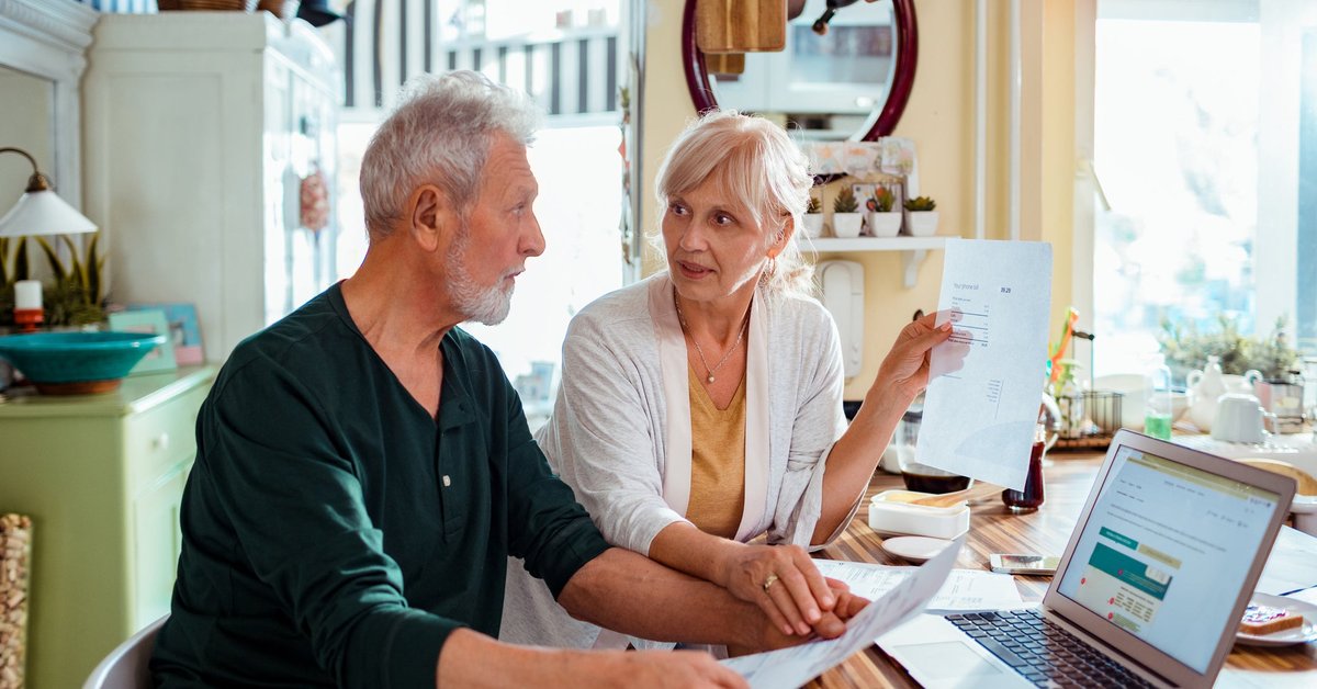 An older couple pays bills at the kitchen table using a laptop.
