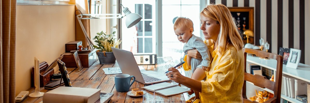 A young parent works on a laptop from home while holding a baby in their lap.