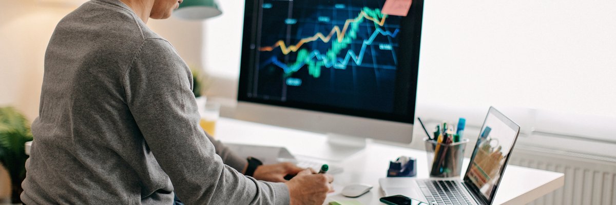 A person using a wheelchair analyzes a stock market graph on their desktop computer and takes notes.