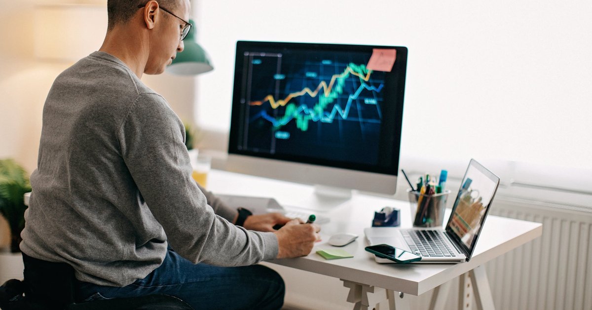 A person using a wheelchair analyzes a stock market graph on their desktop computer and takes notes.