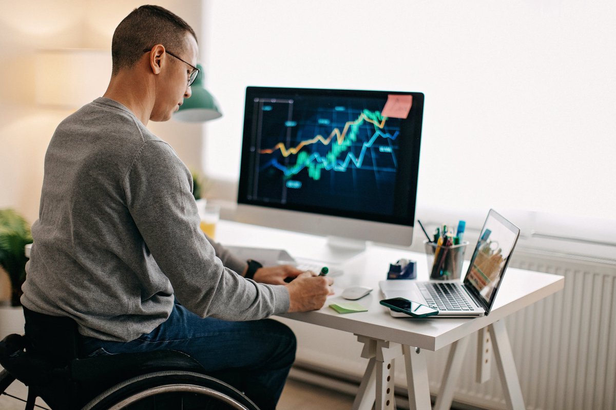 A person using a wheelchair analyzes a stock market graph on their desktop computer and takes notes.