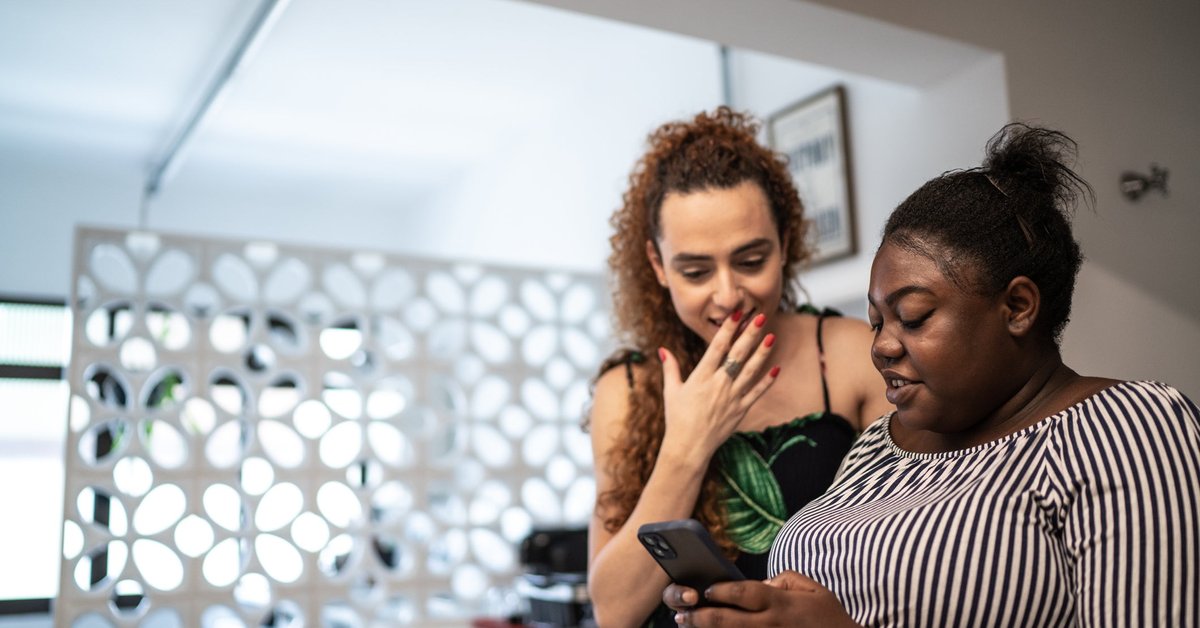 Two friends discuss what's on one of their smartphones in an apartment.