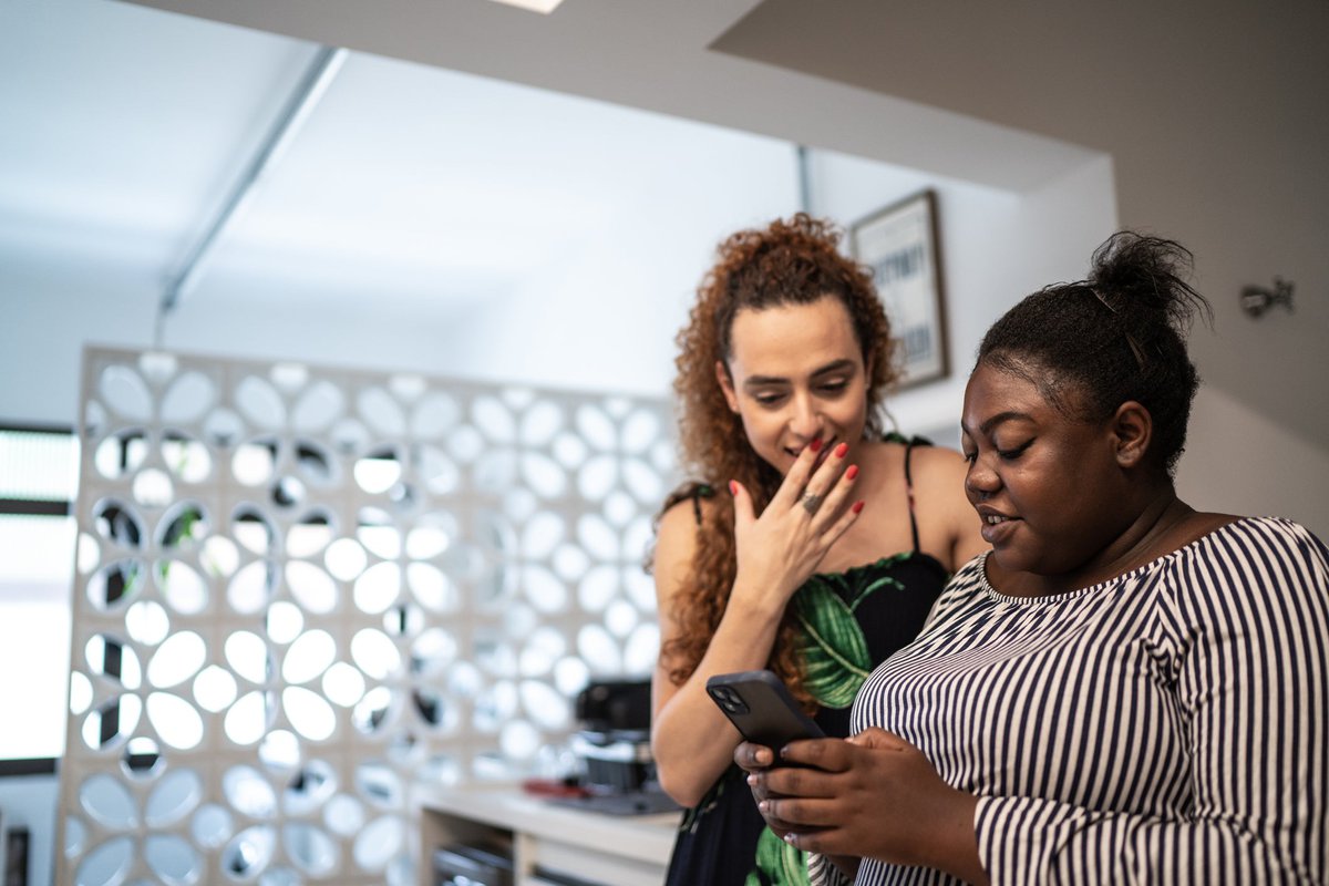 Two friends discuss what's on one of their smartphones in an apartment.