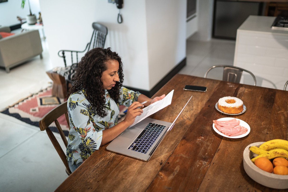 A person looks at financial documents in their kitchen near their laptop.