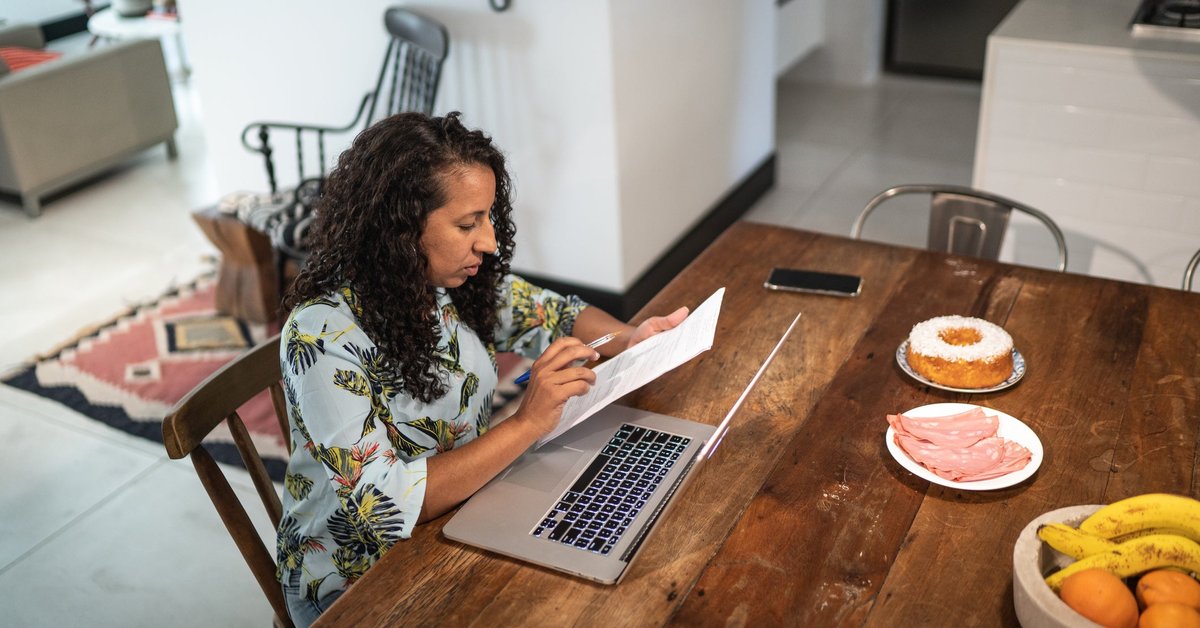 A person manages their home finances using documents and a laptop at their kitchen table.