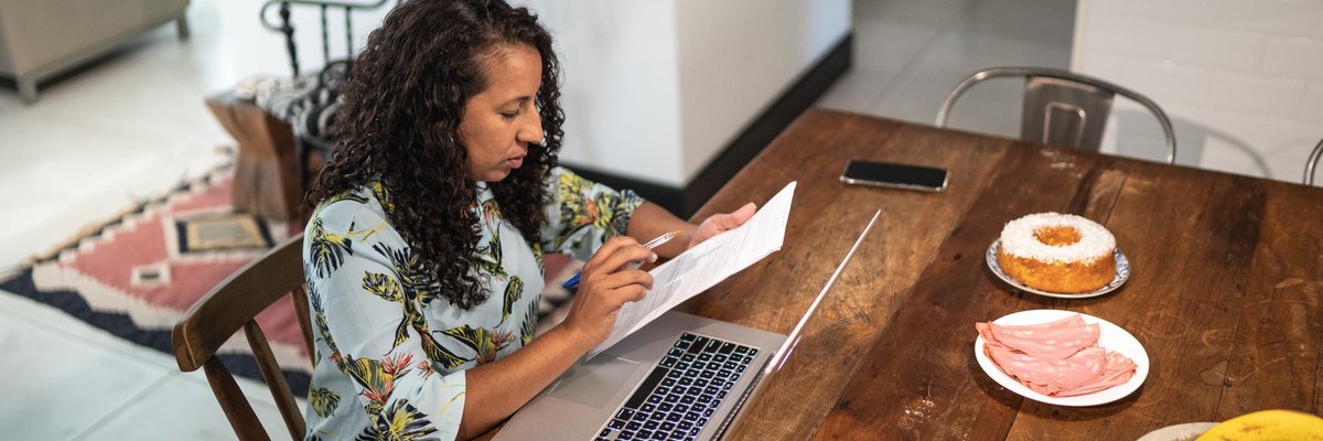 A person manages their home finances using documents and a laptop at their kitchen table.
