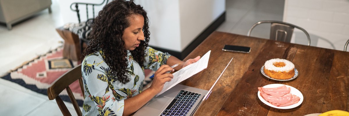 A person manages their home finances using documents and a laptop at their kitchen table.