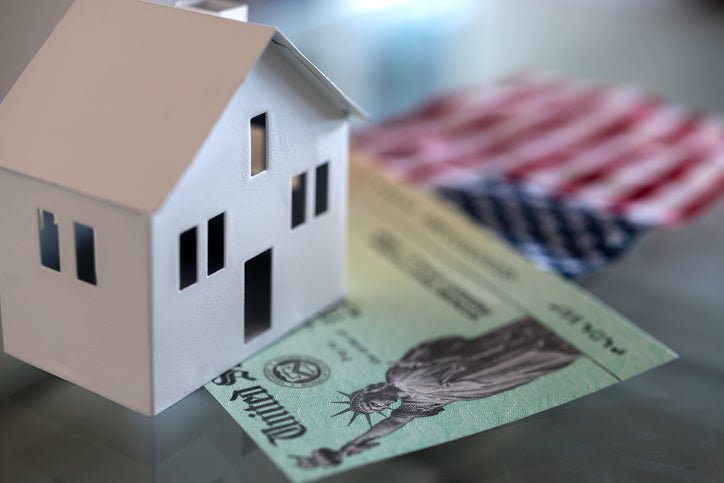 A white cardboard house sits on top of a stimulus check and a small American flag.