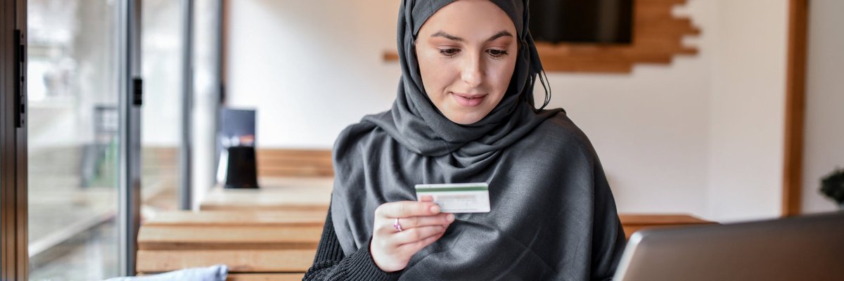 A customer wearing a hijab sits in a coffee shop with a credit card and laptop.