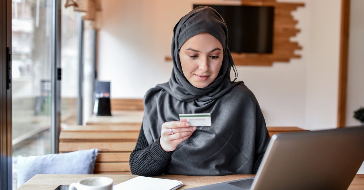 A customer wearing a hijab sits in a coffee shop with a credit card and laptop.