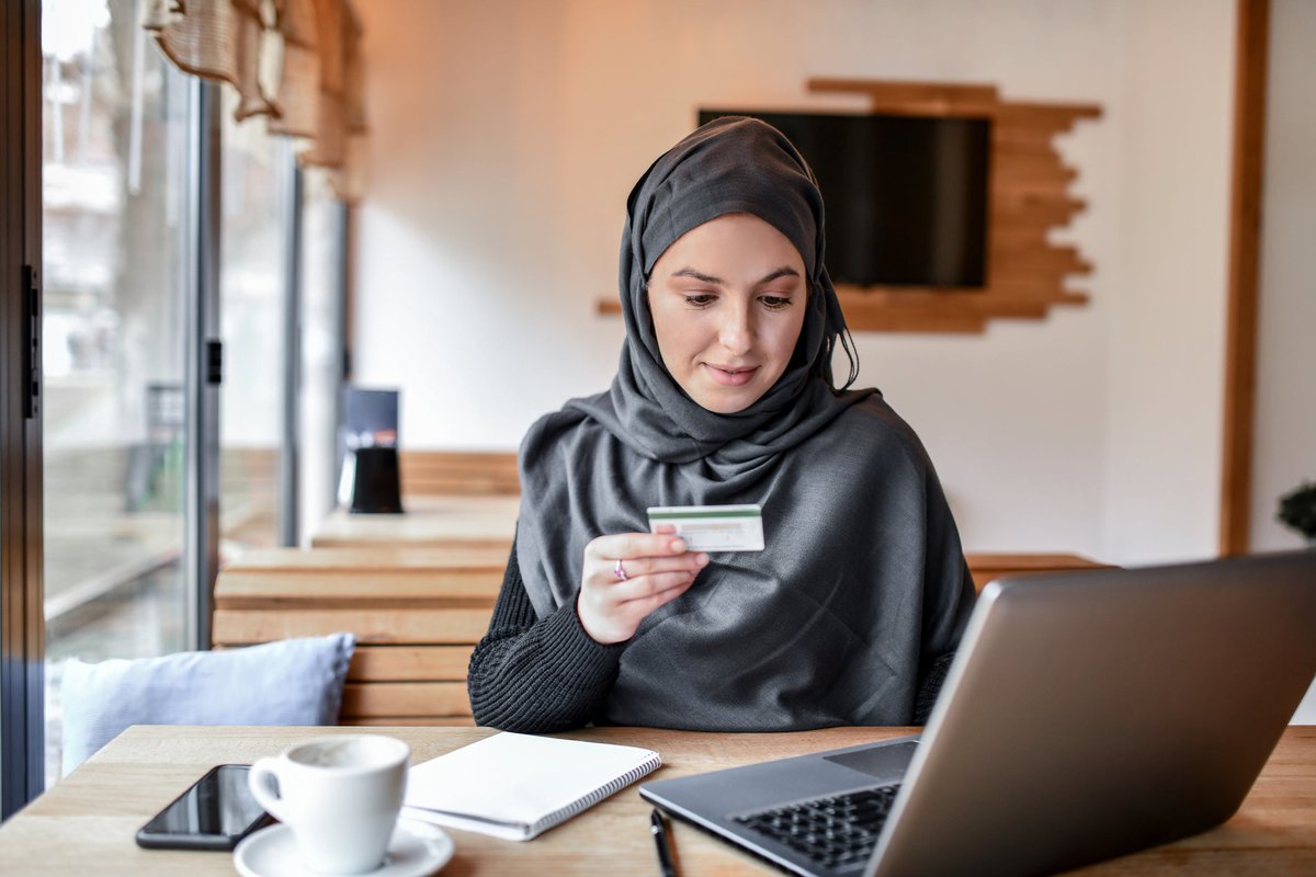 Female customer wearing hijab sits in cafe with credit card and laptop.