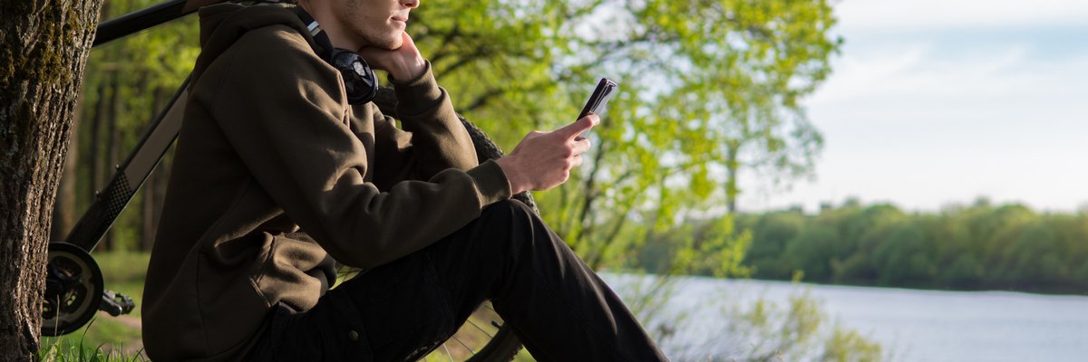 A person sits on a riverbank next to their bicycle, looking at their smartphone.