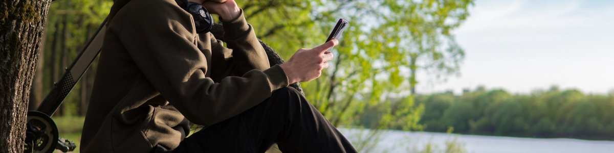 A person sits on a riverbank next to their bicycle, looking at their smartphone.