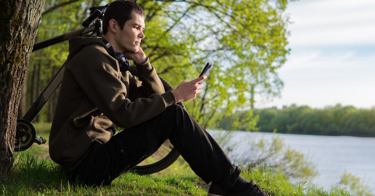A person sits on a riverbank next to their bicycle, looking at their smartphone.