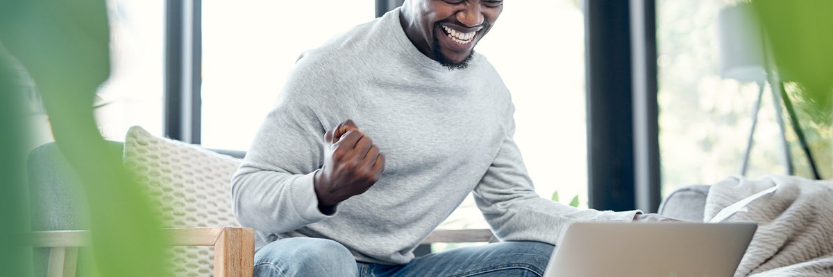 A young adult pumps their fist in celebration while going over paperwork and using a laptop at home.