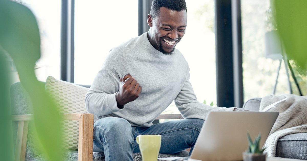 A young adult pumps their fist in celebration while going over paperwork and using a laptop at home.