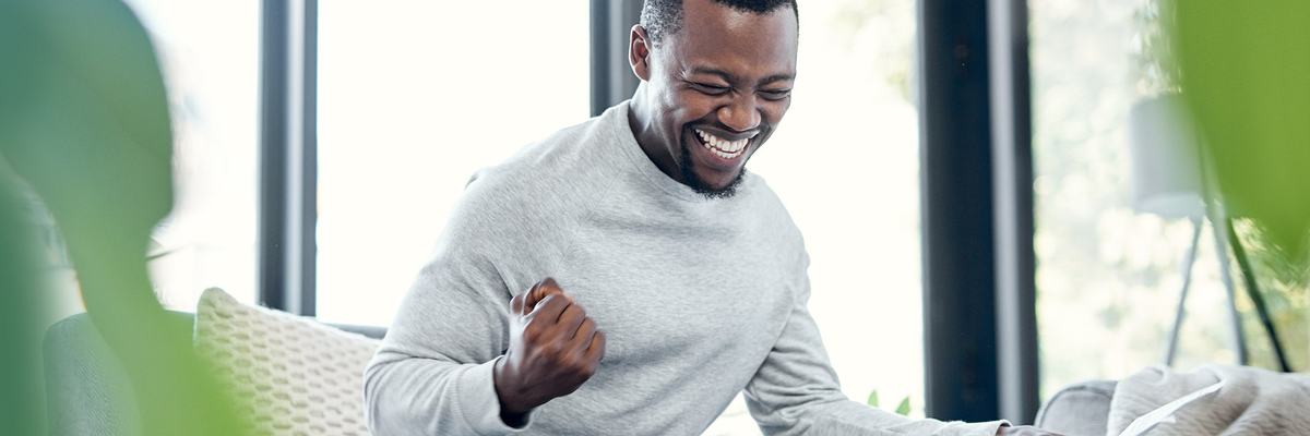 A young adult pumps their fist in celebration while going over paperwork and using a laptop at home.