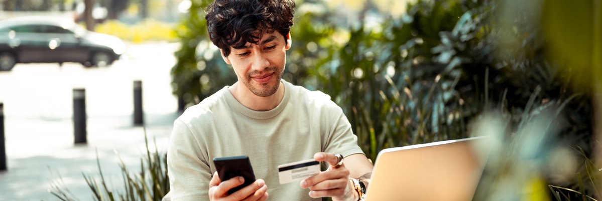 Person sitting outdoors holding credit card and cellphone while using laptop.