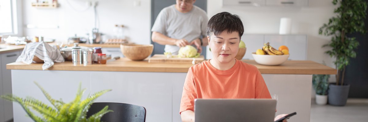 One spouse uses a laptop at the kitchen table while another prepares food in the background.