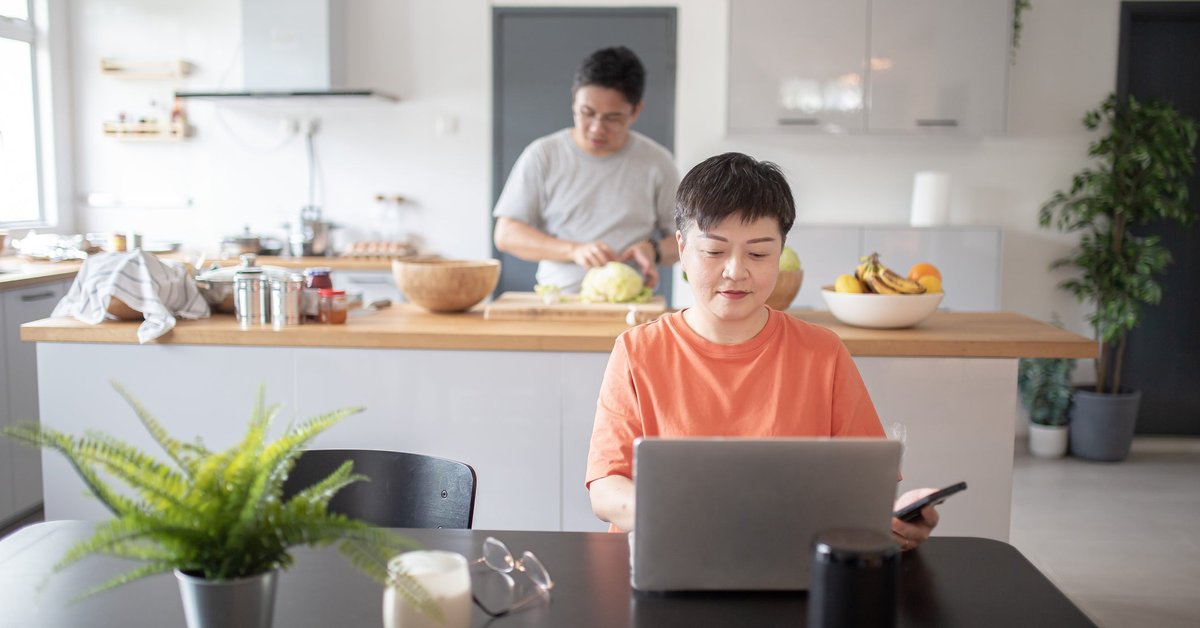 One spouse uses a laptop at the kitchen table while another prepares food in the background.