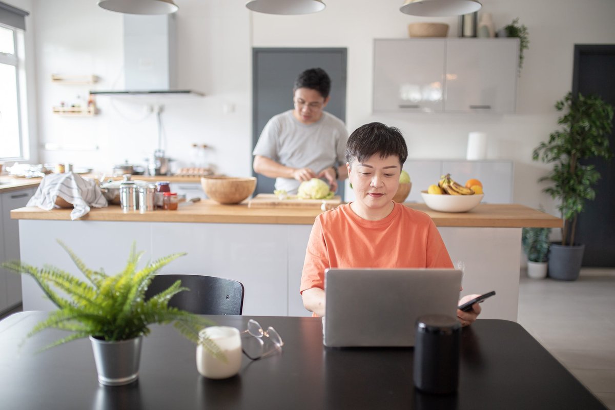 Un conjoint utilise un ordinateur portable à la table de la cuisine tandis qu'un autre prépare la nourriture en arrière-plan.