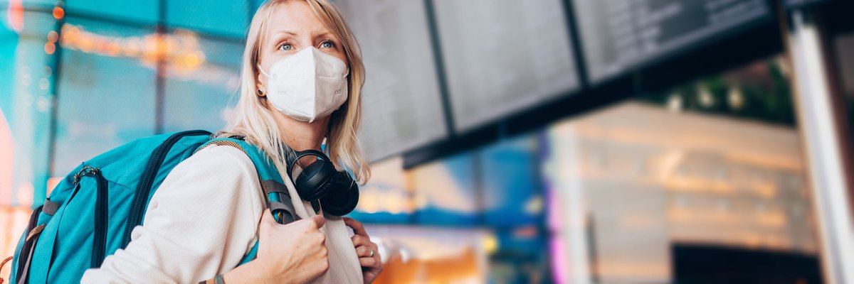A young adult wearing a face mask and a backpack checks the arrivals and departures board at the airport.