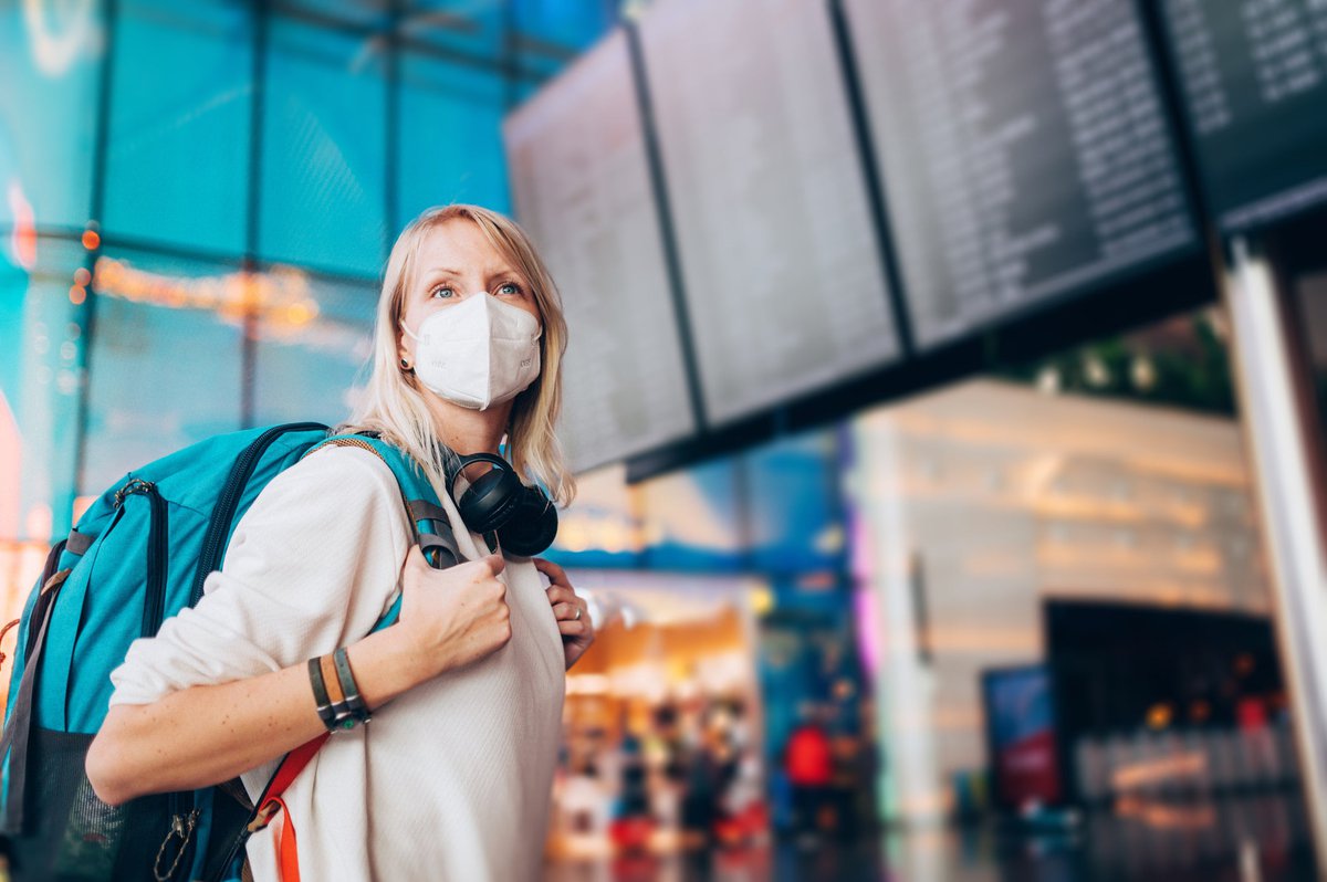 A young adult wearing a face mask and a backpack checks the arrivals and departures board at the airport.