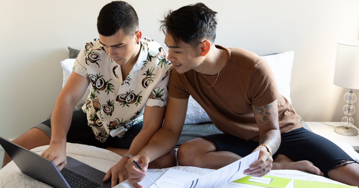 A couple sits on their bed looking at a laptop and paperwork.