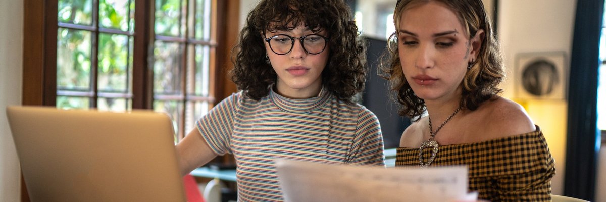 A couple discusses finances at the kitchen table using a laptop and paperwork.