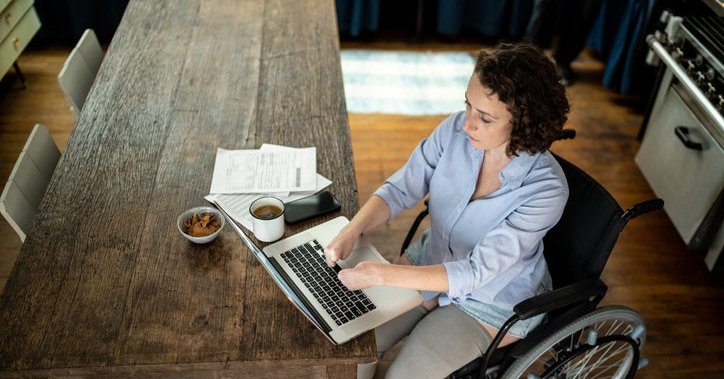 A person using a wheelchair types on their laptop at the kitchen table.