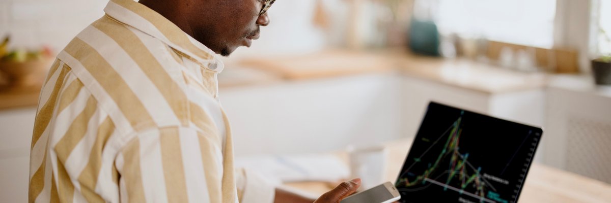 A person analyzes a cryptocurrency graph on their laptop while looking at other information on their smartphone in their kitchen.