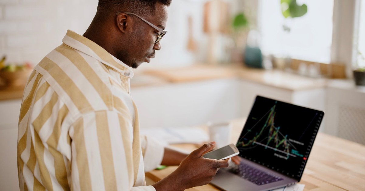 A person analyzes a cryptocurrency graph on their laptop while looking at other information on their smartphone in their kitchen.
