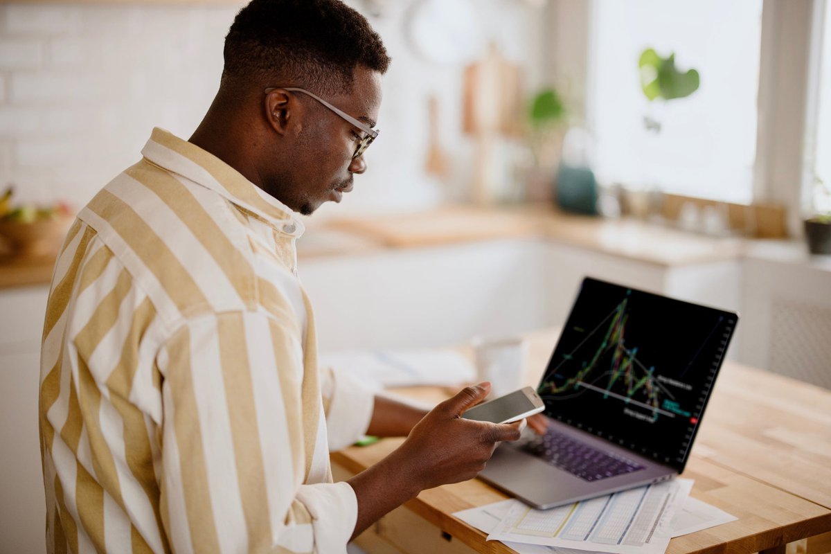 A person analyzes a cryptocurrency graph on their laptop while looking at other information on their smartphone in their kitchen.