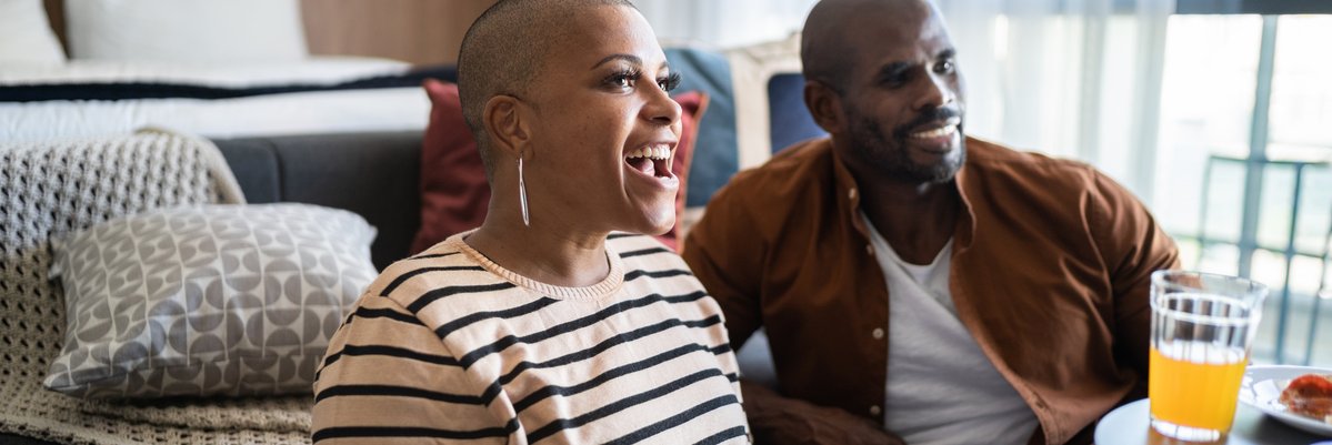 Couple eating pizza and smiling while appearing to watch something on tv.