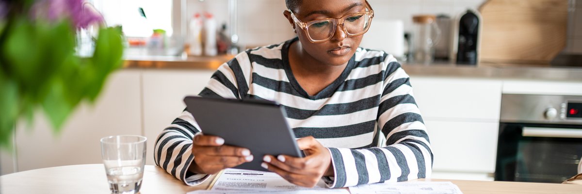 A young adult calculates their personal finances at the kitchen table using a tablet.