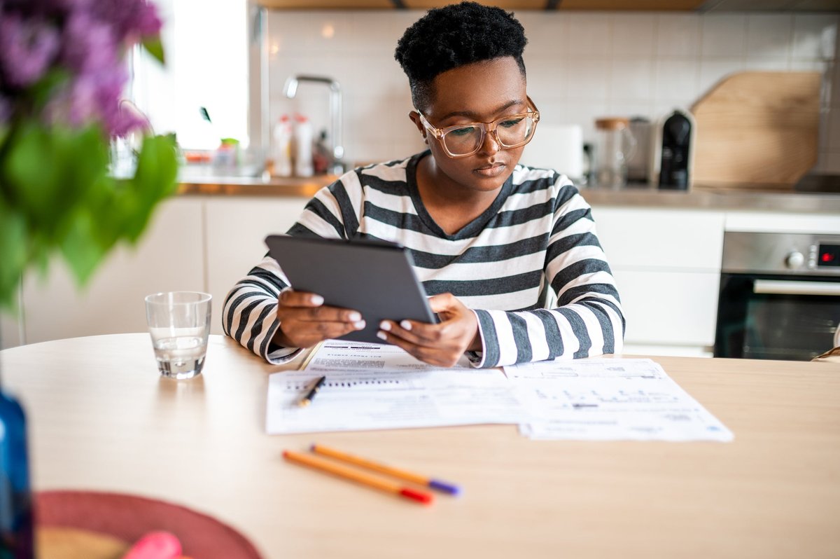 A young adult calculates their personal finances at the kitchen table using a tablet.