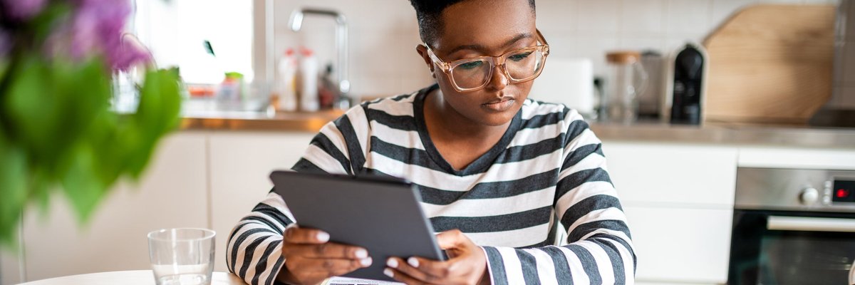 A young adult calculates their personal finances at the kitchen table using a tablet.