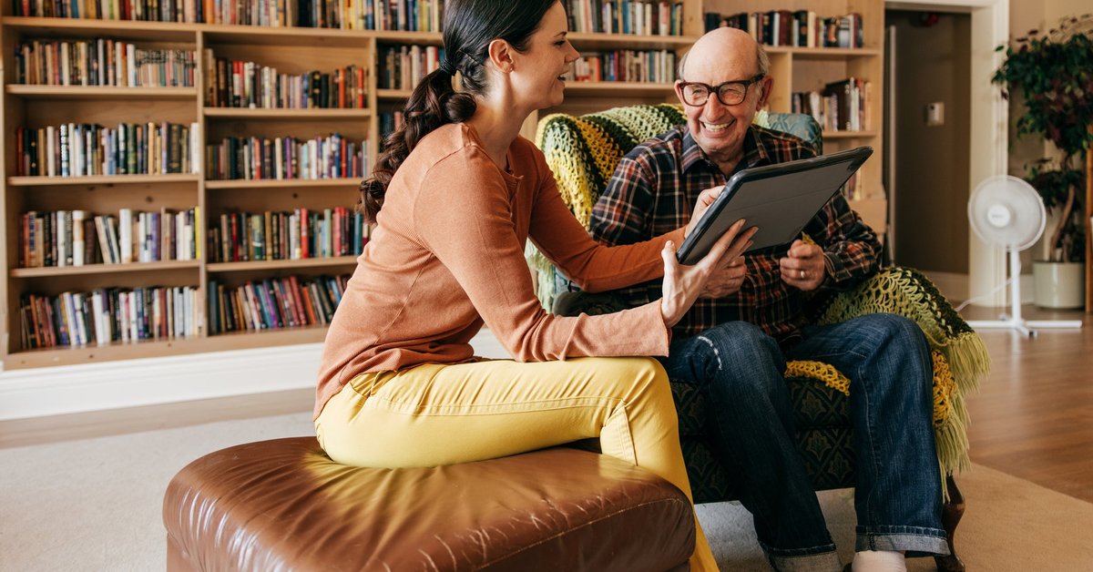 A caregiver holds up a tablet and laughs with their older parent.
