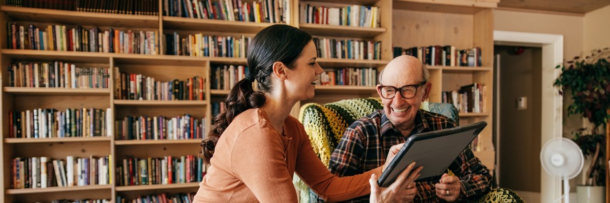 A caregiver holds up a tablet and laughs with their older parent.