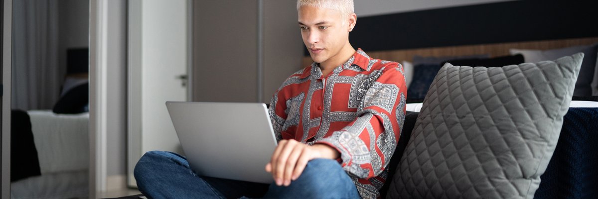 A young adult sits cross-legged while typing on a laptop in their bedroom.