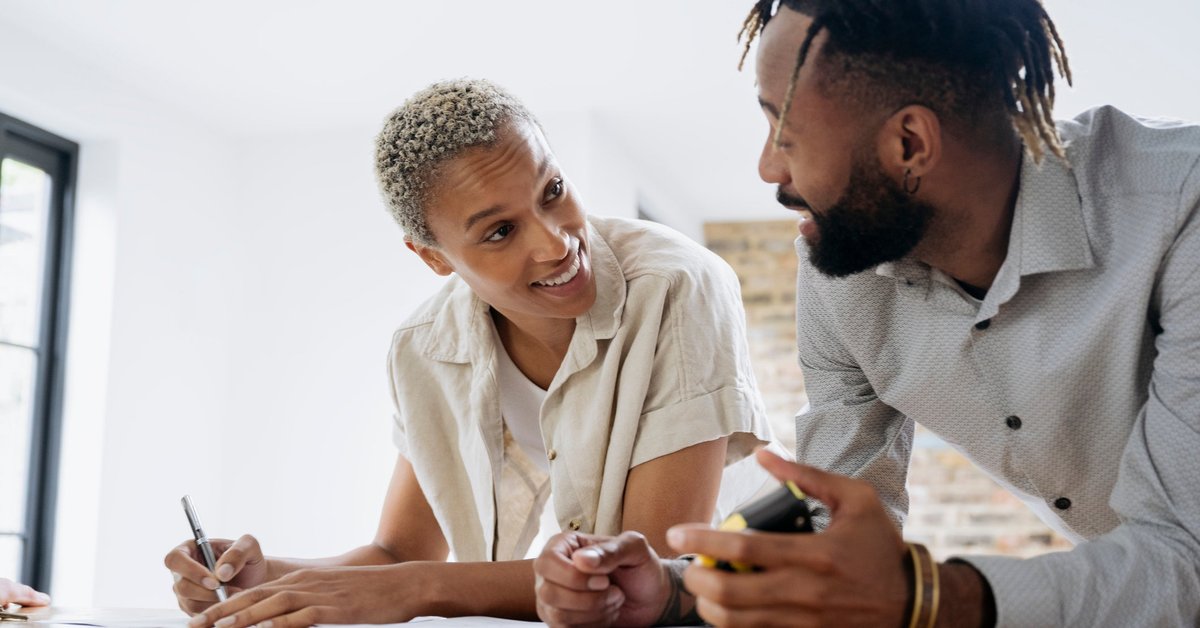 A couple fills out financial paperwork as they smile while leaning against a kitchen island.