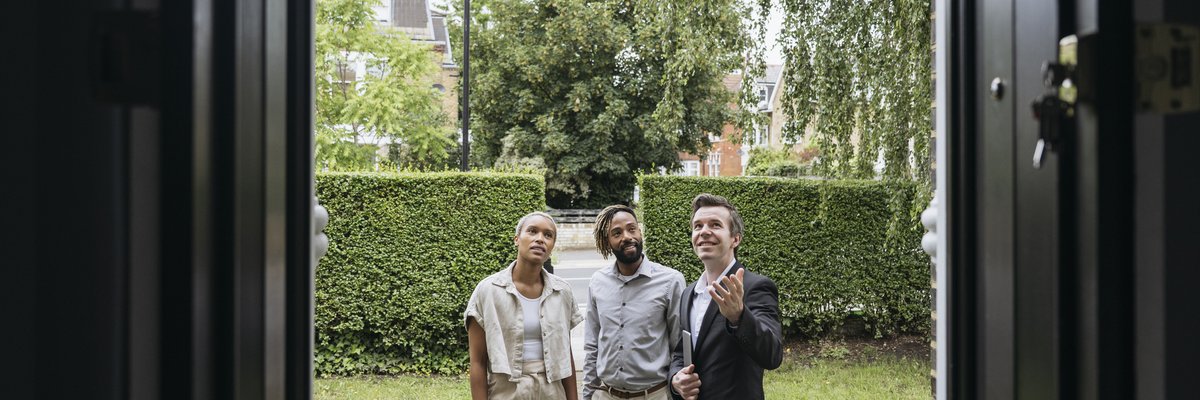 A smiling real estate agent shows two clients features of a home's exterior.