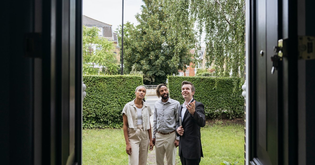 A smiling real estate agent shows two clients features of a home's exterior.