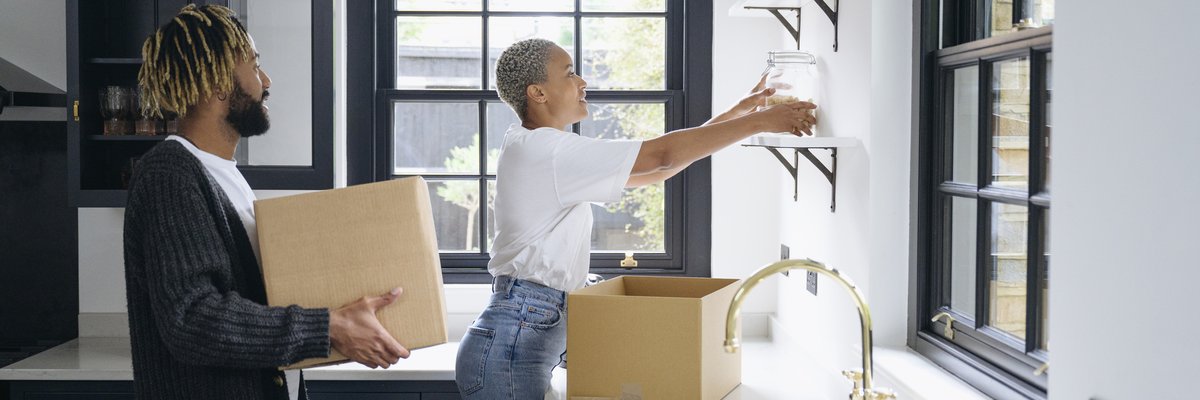 A couple unpacks cardboard boxes and organizes jars on shelves in their new kitchen.