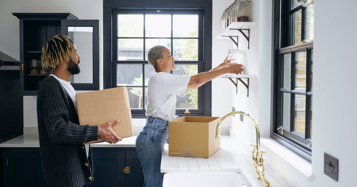 A couple unpacks cardboard boxes and organizes jars on shelves in their new kitchen.