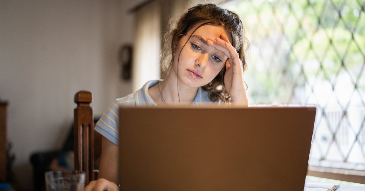 A worried young adult uses a laptop at home.