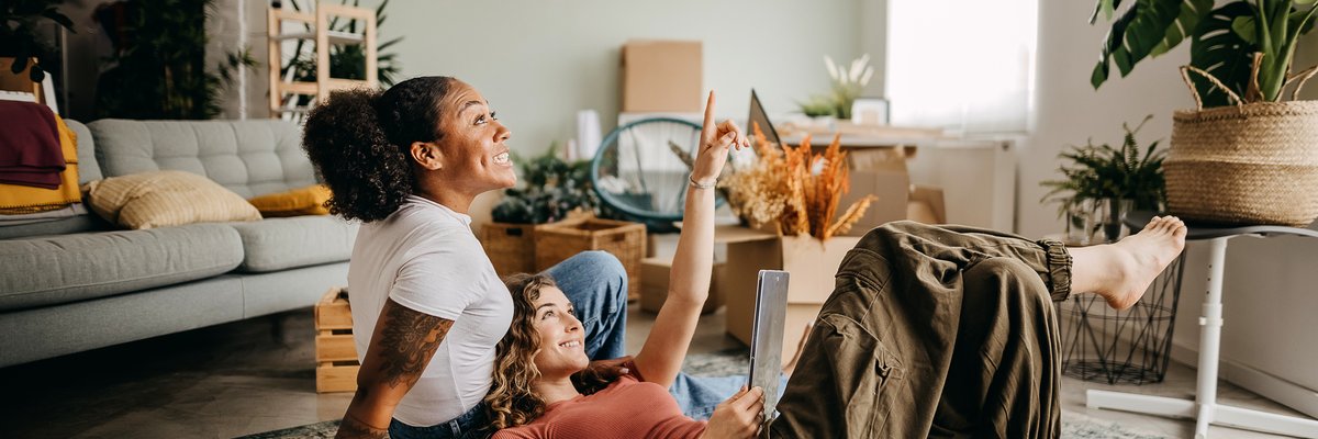 A couple smiles looking at a tablet while sitting on the floor of their apartment.