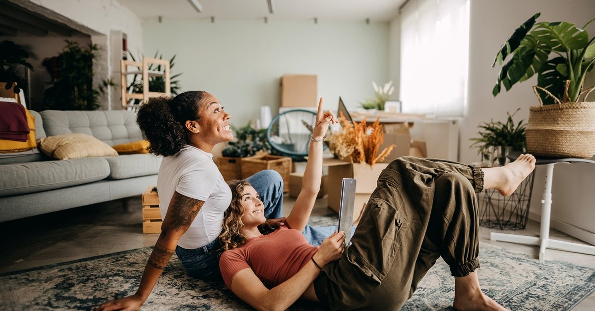 A couple smiles looking at a tablet while sitting on the floor of their apartment.